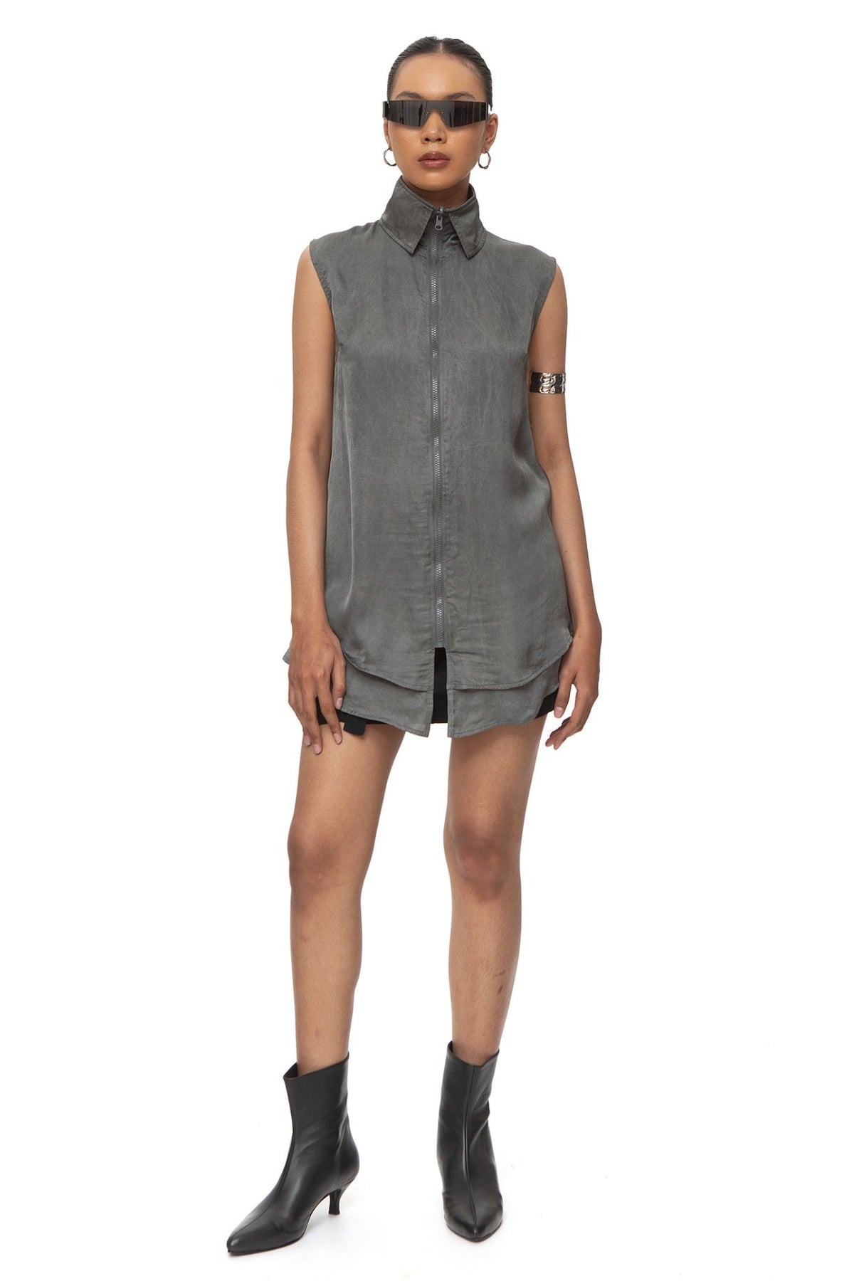 Model wearing grey sleeveless tunic shirt with zip collar and layered curved hem, styled over black shorts